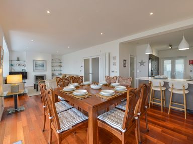 A dining area with a wooden table set for eight and cushioned chairs next to a kitchen with bar stools at Rock Rose in Downderry