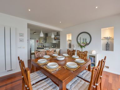 A dining area with a wooden table set for eight and a kitchen in the background at Rock Rose in Downderry