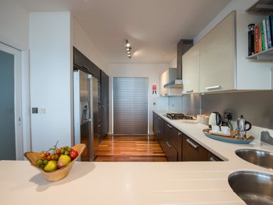 A kitchen with dark lower cabinets and light upper cabinets with a stainless steel refrigerator a gas stove a double sink a wooden bowl with fruit on a white countertop and a shelf with books at Rock Rose in Downderry