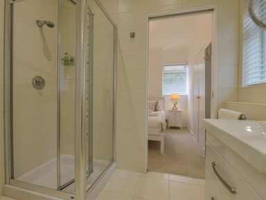 A bathroom with a glass shower enclosure sink and view into a bedroom with a bed lamp and window blinds at Rock Rose in Downderry