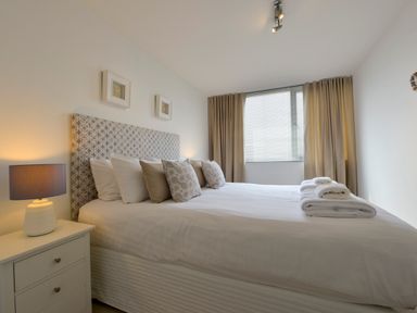 A bedroom with a bed with multiple pillows and folded towels on top beside a nightstand with a lamp and a window with closed blinds at Rock Rose in Downderry