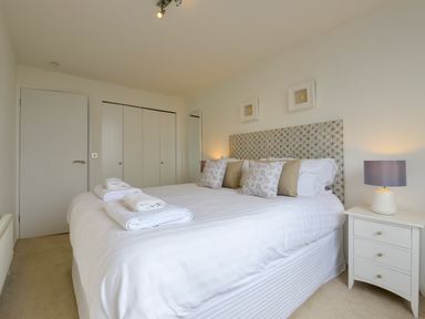 A bedroom with a large bed with white bedding and pillows beside a white nightstand with a lamp at Rock Rose in Downderry