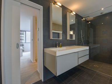 A modern bathroom with a double sink vanity two mirrors a glass shower enclosure and an open door leading to a bedroom at Rock Rose in Downderry