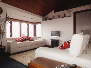 A sitting area with white sofas and Union Jack pillows near windows at Seagulls Nest Whitsand Bay near Millbrook