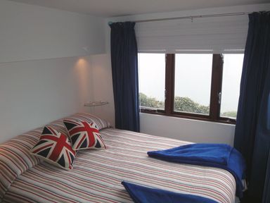 A bedroom with a bed with striped bedding pillows with union jack designs and blue curtains at Seagulls Nest in Whitsand Bay near Millbrook