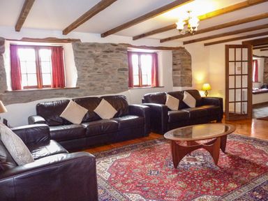 A sitting room with three black leather sofas a red patterned rug a wooden coffee table and exposed stone walls at Samuels Cottage in Lanreath