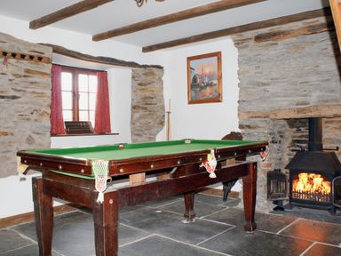 A room with a snooker table a stone fireplace with a wood burning stove a window with red curtains and a painting on the wall at Samuels Cottage in Lanreath