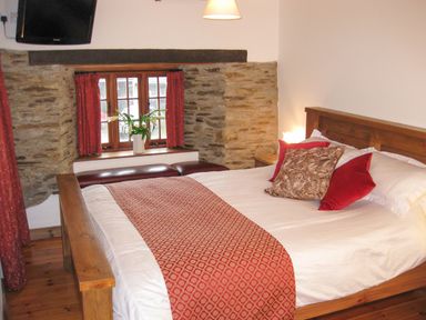A bedroom with a wooden bed, red and patterned pillows, floral curtains, a window with flowers, a wall-mounted TV, and a wooden floor at Samuels Cottage Lanreath