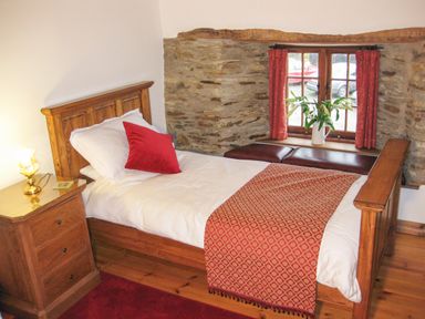 A bedroom with a wooden bed white bedding red pillow and blanket a wooden nightstand with a lamp and a window with red curtains and vase of flowers at Samuels Cottage in Lanreath