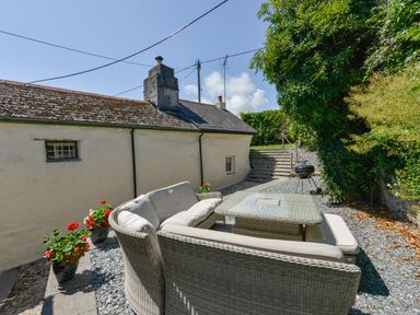 An outdoor patio with wicker seating a table a charcoal grill potted flowers and steps up to a garden at Samuels Cottage in Lanreath