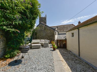 An outdoor patio area with a charcoal grill seating arrangement and potted plants next to stone buildings at Samuels Cottage in Lanreath