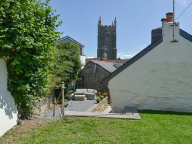 An outdoor seating area with garden furniture and a barbecue surrounded by stone buildings and greenery with a church tower in the background at Samuels Cottage in Lanreath