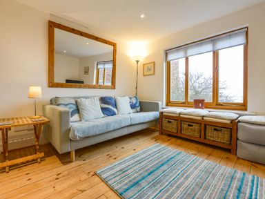 A living room with a sofa cushions a large mirror a wooden side table lamps a window seat with baskets and a striped rug at Chough Cottage in Kingsand And Cawsand
