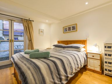 A bedroom with a double bed with striped bedding two rolled towels bedside table with lamp and glass doors with curtains at Chough Cottage in Kingsand And Cawsand