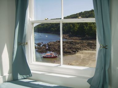 A window with blue curtains looking out over a rocky beach and a red boat at Balcony Cottage in Kingsand And Cawsand