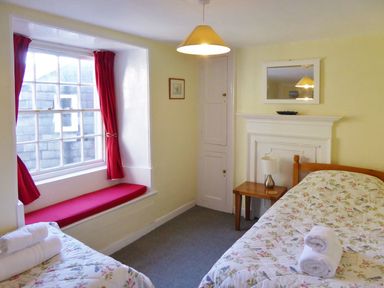 A bedroom with two single beds a window seat with red cushions red curtains and a bedside table with lamp at Balcony Cottage in Kingsand And Cawsand