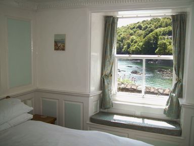 A bedroom with a bed pillow wooden side table window with curtains and seascape view at Balcony Cottage in Kingsand And Cawsand