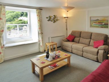 A living room with a beige sofa and red cushions a wooden coffee table with cups and a teapot and a window showing greenery outside at Balcony Cottage in Kingsand And Cawsand