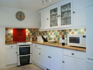 A kitchen with white cabinets and wooden countertops a stove with a red backsplash a microwave a toaster and utensils at Balcony Cottage in Kingsand And Cawsand