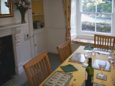 A dining area with a table set with placemats glassware and a bottle of wine near a window with a view of greenery and water at Balcony Cottage in Kingsand And Cawsand