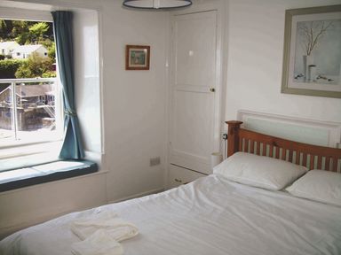 A bedroom with a wooden bed frame white bedding a window seat with cushions and framed pictures on the walls at Balcony Cottage in Kingsand And Cawsand