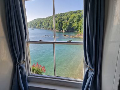 A window with blue curtains showing a view of a body of water and green trees at Balcony Cottage in Kingsand And Cawsand