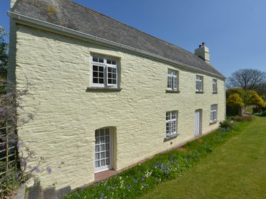 An exterior view of a two-story stone farmhouse with multiple white-framed windows and a garden with grass and flowers at Tregonhawke Farmhouse in Millbrook