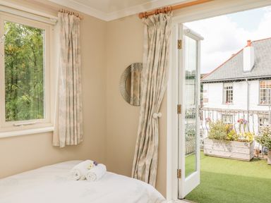 A bedroom with a bed with rolled towels on it next to a window and an open door leading to a balcony with plants at 1 River Cottage in Calstock