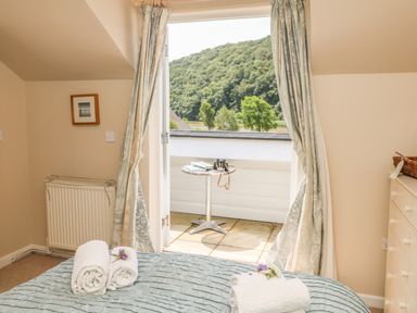 A bedroom with a bed and rolled towels near an open door leading to a balcony with a table at 1 River Cottage in Calstock