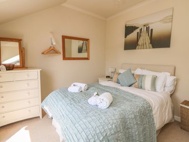 A bedroom with a bed covered in a blue bedspread and rolled towels on top near a white dresser and a wall mirror at 1 River Cottage in Calstock