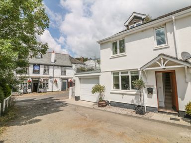 An exterior view of a white house with open door and garage next to a white building with hanging flower baskets and sign at 1 River Cottage in Calstock