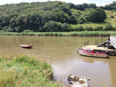 A river with three boats near a grassy bank and a dock with railings with trees and hills in the background at 1 River Cottage in Calstock