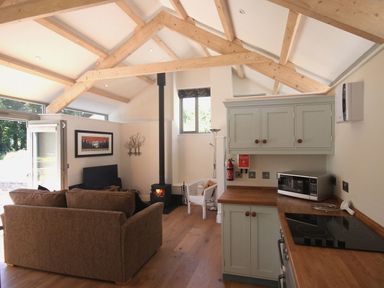 An open plan living room and kitchen with wooden ceiling beams a stove a sofa and kitchen cabinets at Mugwell Barn Luckett near Callington