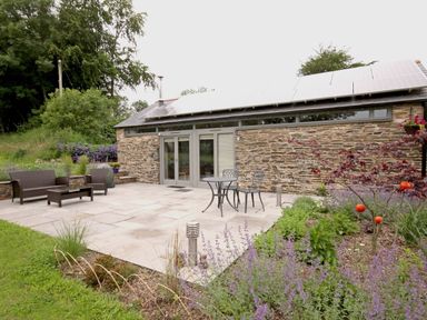 An outdoor patio with stone tiles a table and chairs a wicker bench and chair plants and solar panels on roof at Mugwell Barn Luckett near Callington