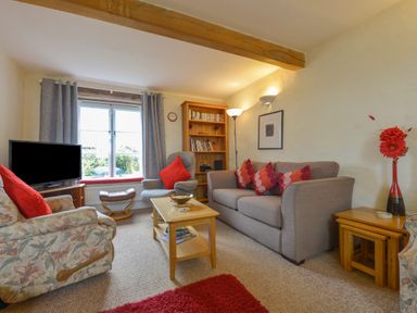 A living room with a gray sofa and armchairs around a wooden coffee table with a bookshelf and window in the background at Regatta Cottage in Feock