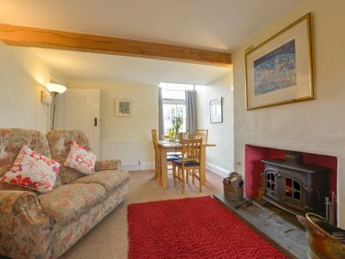 A living room with a floral sofa two pillows a red rug a wood burning stove and a dining table with chairs at Regatta Cottage in Feock