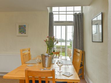 A dining room with a wooden table and chairs set with placemats and glasses near a window with grey curtains at Regatta Cottage in Feock