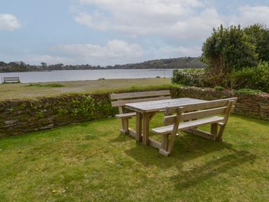 A wooden picnic table with benches on a lawn near a low stone wall overlooking a body of water with hills and trees in the background at Regatta Cottage in Feock