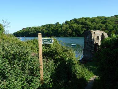A wooden public footpath sign near bushes by a water body with a stone structure and trees in the background at Regatta Cottage in Feock