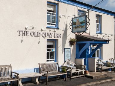 The exterior of The Old Quay Inn with wooden benches and a sign featuring a sailing boat