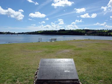 A grassy area with a bench and a metal plaque near a body of water with hills in the background at Regatta Cottage in Feock