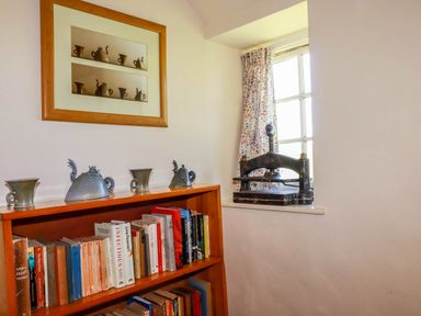 A corner of a room with a wooden bookshelf holding books and pottery with a window and an old press on the windowsill at Polcreek Farmhouse in Veryan