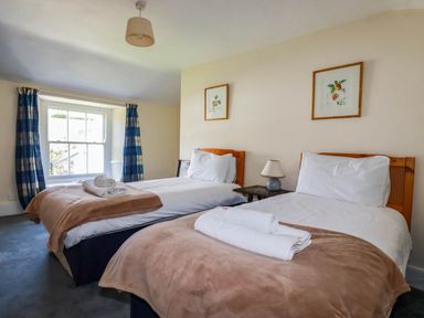 A bedroom with two single beds with white linens and beige blankets a window with blue and white curtains and framed pictures on the wall at Polcreek Farmhouse in Veryan