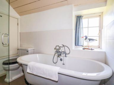 A bathroom with a bathtub a toilet a window with a curtain and a bird decoration at Polcreek Farmhouse in Veryan