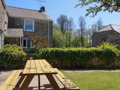 A wooden picnic table on a grass and paved area outside stone buildings with greenery and trees in the background at Polcreek Farmhouse in Veryan