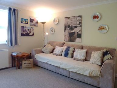 A living room with a sofa with cushions by a window with curtains and framed pictures on the wall at Stones Throw in St Mawes