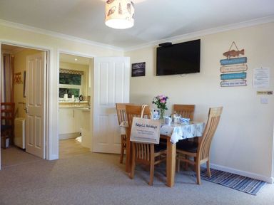 A dining area with a wooden table and chairs a wall mounted television and an open door to a kitchen at Stones Throw in St Mawes