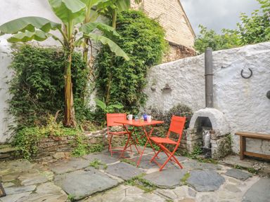 An outdoor patio with orange table and chairs surrounded by plants and a white wall with a small fireplace and a bench at Primrose Cottage Penryn