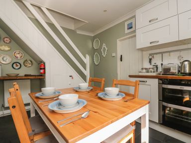 A kitchen with a wooden dining table set for four under a staircase with decorative plates on the wall at Primrose Cottage in Penryn
