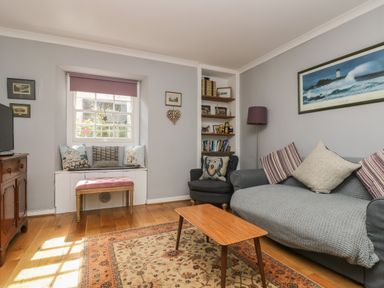A living room with a grey sofa and armchair a wooden coffee table and a window seat with cushions at Primrose Cottage in Penryn